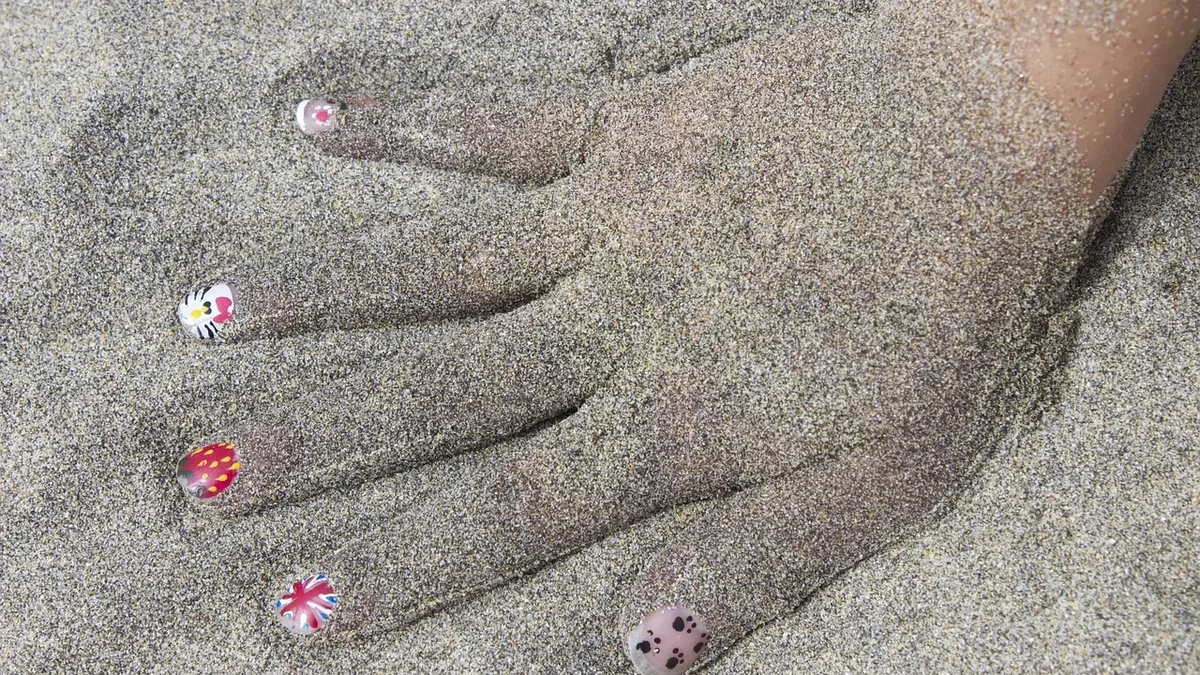 A hand with painted fingernails, partially buried in sand.
